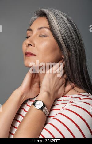 Immagine di donna stanca matura adulto che indossa camicia a righe toccando il collo isolato su sfondo grigio Foto Stock