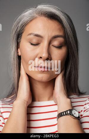 Immagine di donna stanca matura adulto che indossa camicia a righe toccando il collo isolato su sfondo grigio Foto Stock