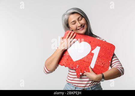 Immagine di donna felice adulto matura con lunghi capelli bianchi che tengono il cuore come simbolo sul placard isolato su sfondo grigio Foto Stock