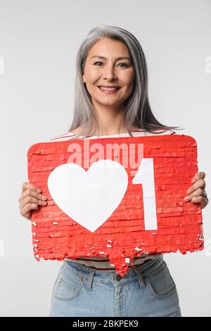 Immagine di donna felice adulto matura con lunghi capelli bianchi che tengono il cuore come simbolo sul placard isolato su sfondo grigio Foto Stock