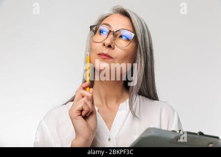 Ritratto di un'attraente donna d'affari matura e penosa in abiti formali in piedi isolato su sfondo bianco, tenendo un clipboard Foto Stock