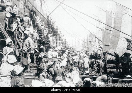 [ 1910 Giappone - Caricamento del carbone a Nagasaki ] – una folla di portuali carica carbone di alta qualità Kyushu su una nave a Nagasaki. Non appena le navi entrarono nel porto di Nagasaki, centinaia di uomini, donne, ragazze e ragazzi si sciolse intorno a loro su chiatte di carbone, costruirono strutture temporanee e iniziarono a caricare le navi affamate di carbone. stampa d'argento gelatina vintage del xx secolo. Foto Stock