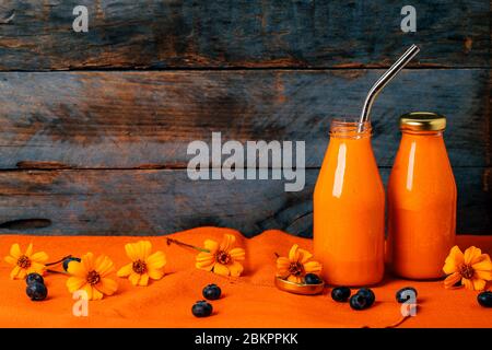 Frullato di arance e carote in bottiglie di vetro con paglia inossidabile con fiori di Chernobrivtsi e mirtilli su fondo rustico di legno grunge con stoffa. Foto Stock