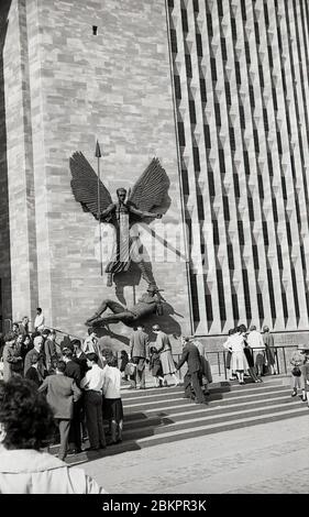 1961, immagine storica della gente fuori della nuova cattedrale, Coventry, Inghilterra alla sua apertura. La scultura sul muro, di Jacob Epstein, denota la vittoria di San Michele sul Diavolo. Pesantemente bombardato durante la seconda guerra mondiale, nel maggio 1962 fu ufficialmente aperto un nuovo edificio 'modernista' sul sito delle rovine della vecchia cattedrale. L'edificio fu progettato dalla Basilio Spence nel radicale stile architettonico 'Brutalista', comune in questa epoca. Foto Stock