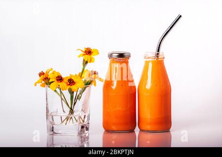 Frullato di arance e carote in bottiglie di vetro con coperchio in acciaio inox e paglia, e vetro con fiori di Chernobrivtsi su fondo bianco. Foto Stock