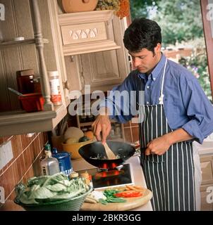 Anni '90 uomo 25-30 anni di cucina una sana agitazione-friggere con piano cottura, nella sua cucina stile anni '90, con giardino soleggiato in background attraverso la finestra della cucina aperta Foto Stock