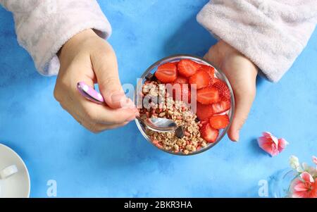 Colazione. Donna mani tiene una ciotola con fragole, cereali e yogurt. Sfondo blu. Vista dall'alto. Foto Stock