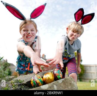 Un fratello e una sorella, indossando la pittura facciale e le orecchie conigliate fatte in casa, prendere parte a una caccia all'uovo di Pasqua la Domenica di Pasqua nel loro giardino. Foto Stock