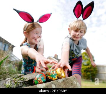 Un fratello e una sorella, indossando la pittura facciale e le orecchie conigliate fatte in casa, prendere parte a una caccia all'uovo di Pasqua la Domenica di Pasqua nel loro giardino. Foto Stock