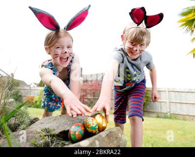 Un fratello e una sorella, indossando la pittura facciale e le orecchie conigliate fatte in casa, prendere parte a una caccia all'uovo di Pasqua la Domenica di Pasqua nel loro giardino. Foto Stock
