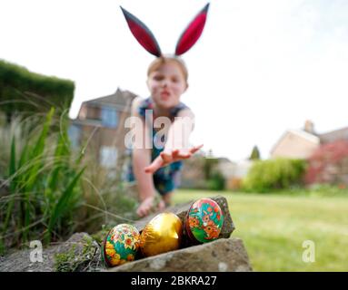 Una giovane ragazza che indossa la vernice del viso e le orecchie conigliate fatte in casa, partecipa a una caccia all'uovo di Pasqua la Domenica di Pasqua nel suo giardino. Foto Stock