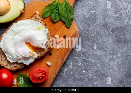 Uovo in camicia su pane con semi, pomodoro di ciliegia, spinaci, avocado, sale e spezie su un vassoio di legno su fondo pietra Foto Stock