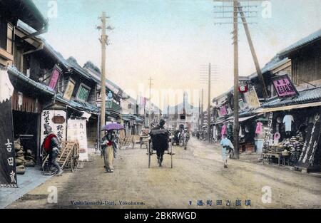 [ 1900 Giappone - Negozi giapponesi a Yokohama ] - UN risciò e una donna con ombrello passano accanto ai negozi su Yoshidamachi-dori, Yokohama, Prefettura di Kanagawa. Il fotografo guardò da 2-chome verso 1-chome. L'edificio sul retro è la stazione di polizia di Isezaki (伊勢佐木警察署, Ikezaki Keisatsusho). Di fronte alla stazione, a sinistra, si trova il ponte Yoshidabashi. cartolina vintage del xx secolo. Foto Stock