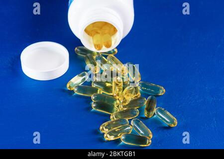 Capsule gialle masticabili con olio di pesce in vaso bianco di plastica sono adagiate su sfondo blu. Omega 3 pillole come supporto per la salute e il concetto di trattamento. Biolo Foto Stock
