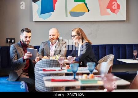 Giovane manager barbuto in giacca utilizzando tablet digitale mentre presenta il progetto aziendale ai colleghi di un moderno caffè Foto Stock