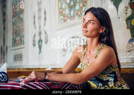 Bella donna brunetta seduta a un tavolo, guardando pensieroso di lato. Sullo sfondo sono decorazioni esotiche; le braccia si appoggiano sul tavolo. Foto Stock