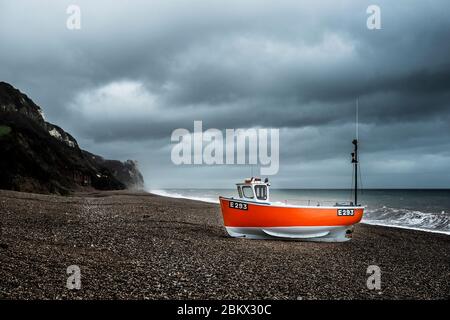 Una barca da pesca a scafo rosso sulla spiaggia di ciottoli a Branscombe, Devon, con un cielo tempestoso Foto Stock