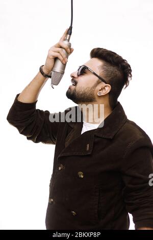 Giovane uomo di talento che indossa un cappotto marrone e un cappotto di occhiali che cantano una canzone in studio di registrazione Foto Stock