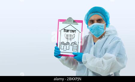 Medico che guarda dritto con maschera e guanti blu tenendo con le mani una tavola con un foglio bianco con messaggio: STARE AL SICURO. Set con guanti, ma