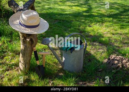 Cappello da sole con guanti, lattina d'irrigazione e attrezzi da giardinaggio adagiato su un prato verde in un giardino primaverile al sole concettuale delle stagioni Foto Stock