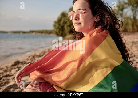 Giovane donna avvolta in bandiera arcobaleno sulla spiaggia Foto Stock