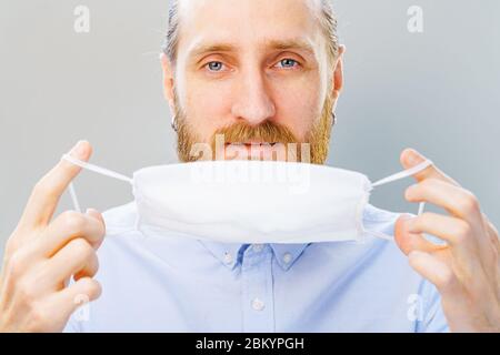 L'uomo bianco bearded in camicia blu mette su una maschera di inquinamento dell'aria e di coronavirus Covid19, guardando la macchina fotografica. Studio girato su sfondo grigio Foto Stock