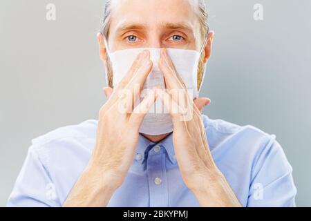 L'uomo bianco bearded in camicia blu mette su una maschera di inquinamento dell'aria e di coronavirus Covid19, guardando la macchina fotografica. Studio girato su sfondo grigio Foto Stock