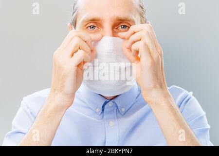 L'uomo bianco bearded in camicia blu mette su una maschera di inquinamento dell'aria e di coronavirus Covid19, guardando la macchina fotografica. Studio girato su sfondo grigio Foto Stock