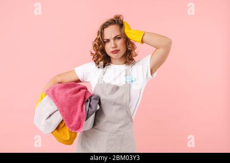 Immagine di una giovane donna confusa stanca casalinga in guanti isolati su sfondo rosa parete che tiene lavanderia. Foto Stock