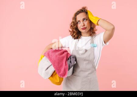 Immagine di una giovane donna confusa stanca casalinga in guanti isolati su sfondo rosa parete che tiene lavanderia. Foto Stock