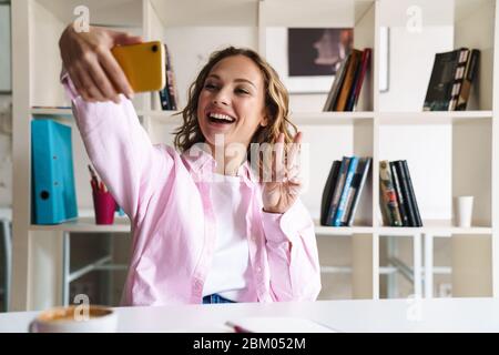 Foto di giovane donna sorridente che prende selfie sul cellulare e gesturing segno di pace in bar Foto Stock