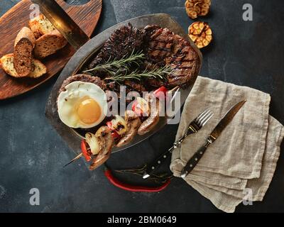 Carne alla griglia assortita, verdure e spezie, un tavolo da cucina per servire la carne, con tovaglioli spalmabili e coltelli. Foto Stock
