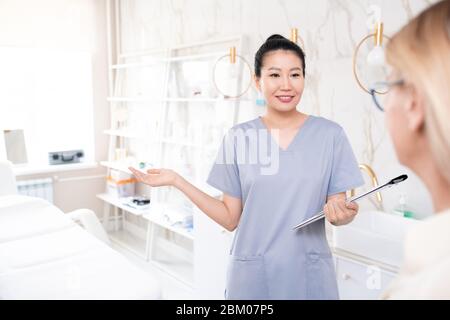 Sorridente cosmetologa carino in scrub tenendo appunti e accogliente cliente in salone di bellezza Foto Stock