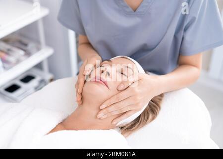 Primo piano di un professionista di bellezza irriconoscibile che offre massaggio facciale a donna matura rilassata nel salone di bellezza Foto Stock