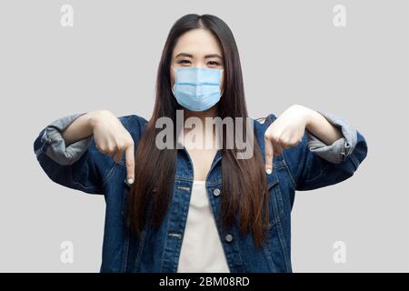 Qui e adesso. Ritratto di bruna grave bossy donna asiatica giovane con maschera medica chirurgica in giacca blu denim in piedi e rivolto verso il basso. S Foto Stock