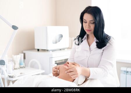 Attraente giovane beautician che dona massaggio viso mano a donna matura in salone di bellezza Foto Stock