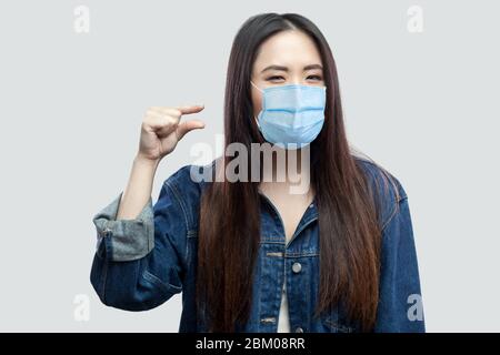 Dammi qualche altro. Ritratto di giovane donna asiatica brunetta con maschera medica chirurgica in giacca casual blu denim in piedi e mostrando un piccolo taglia Wit Foto Stock