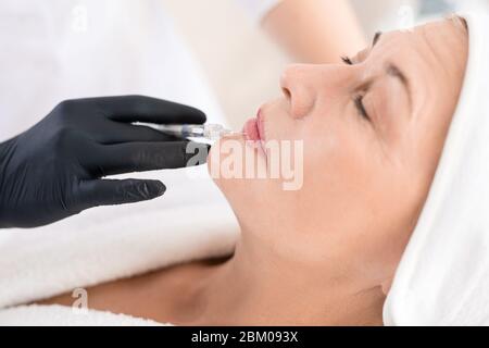 Mano in guanto nero di cosmetologist che fa l'iniezione di aumento del labbro alla donna matura Foto Stock