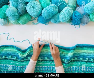 Le mani delle donne sono grandi. Donna crochets. Filati di colore verde, turchese, acquamarina e blu. Sfondo in legno bianco. Foto Stock