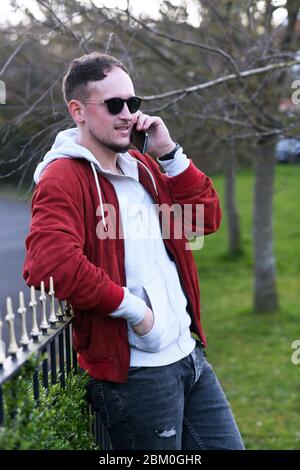 Uomo alla moda e impegnato in abiti casual e occhiali da sole che guardano via e fanno una chiamata mentre si sta in piedi con la mano in tasca e appoggiandosi sul recinto sulla strada Foto Stock