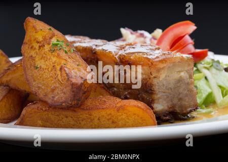 Costolette di maiale con patate arrosto, cavolo e lattuga in un piatto bianco di ceramica su un tavolo di legno Foto Stock
