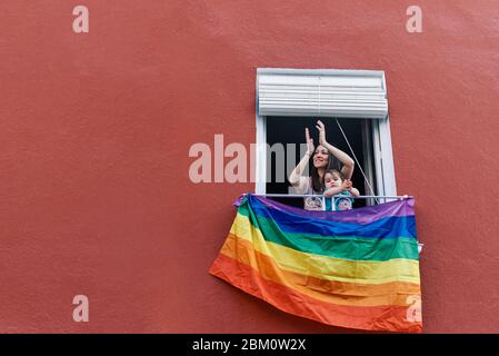 giovane donna in una maschera che si aggrasta dalla finestra con la bandiera gay su uno sfondo rosso Foto Stock