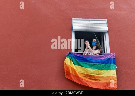 giovane donna in una maschera che si aggrasta dalla finestra con la bandiera gay su uno sfondo rosso Foto Stock