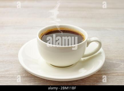 Caffè espresso nero caldo con vapore in tazza bianca, piattino su sfondo rustico tavolo in legno. Foto Stock