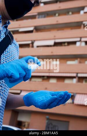 primo piano di un uomo caucasico per strada, indossando una maschera nera per il viso e guanti in lattice blu, disinfettando le mani spruzzando un disinfettante blu da un Foto Stock