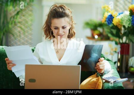 donna moderna in blusa bianca con documenti e calcolatrice scegliendo assicurazione medica digitalmente nella casa moderna in giorno di sole. Foto Stock