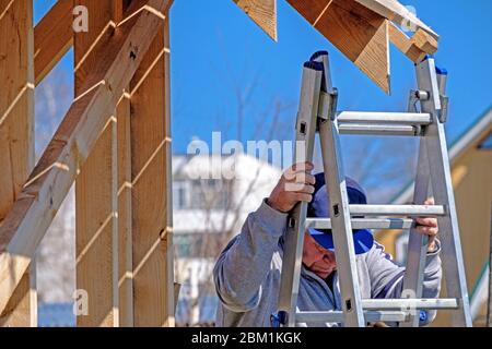 Il costruttore senior grigio-capelli scende le scale contro il cielo blu. Il processo di costruzione di una cornice di una casa di campagna in legno. L'attività fisica Foto Stock