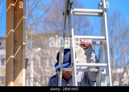 Il costruttore senior grigio-capelli scende le scale contro il cielo blu. Il processo di costruzione di una cornice di una casa di campagna in legno. L'attività fisica Foto Stock