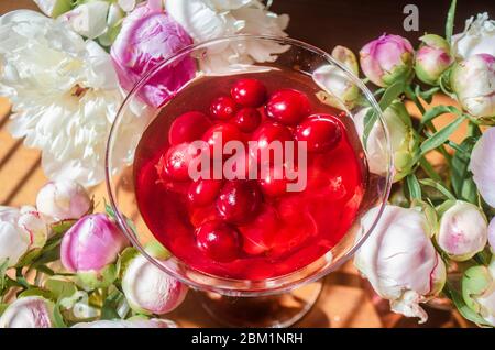 Magia solenne vita morta con bicchiere di bevanda rossa con bacche biologiche autunnali circondate da fiori bianchi nell'ombra diagonale a righe dei ciechi o Foto Stock