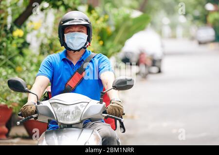 Corriere in casco e maschera medica in sella allo scooter quando si consegna cibo ai clienti che rimangono a casa durante la pandemia Foto Stock